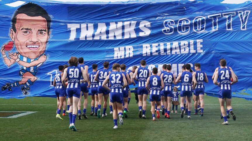 Male AFL players run towards a banner as they run on the field.