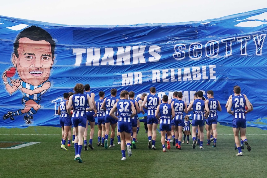 Male AFL players run towards a banner as they run on the field.