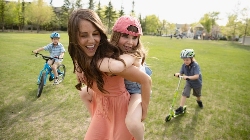 A woman piggybacks a girl in a park, while two children ride bikes in the background.