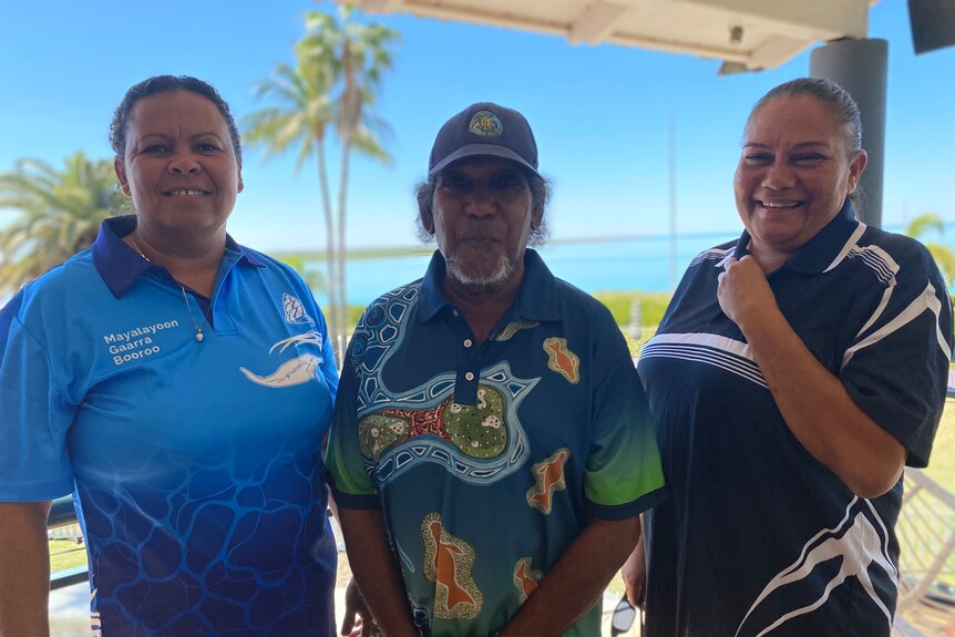A man and two women standing in front of blue water, all with Aboriginal corporation shirts on