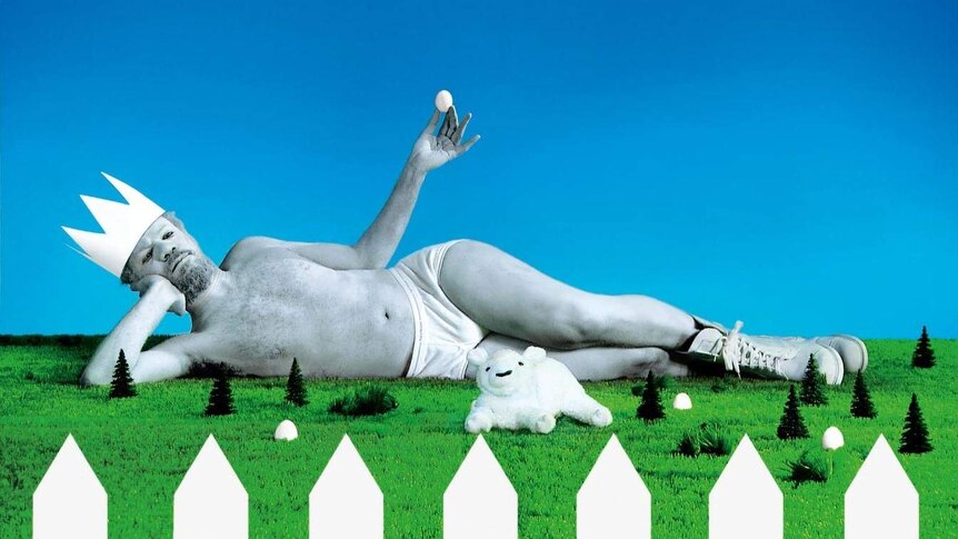A man in white briefs and painted in white stage makeup is lying on his side on fake grass, holding an egg up with one hand
