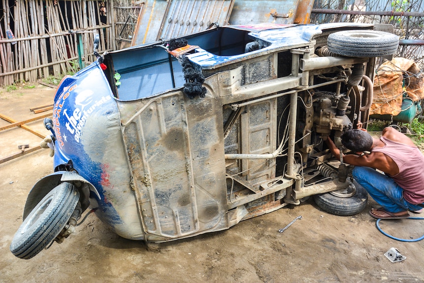 A man tinkers with a tuk-tuk lying on its side on a dusty road.