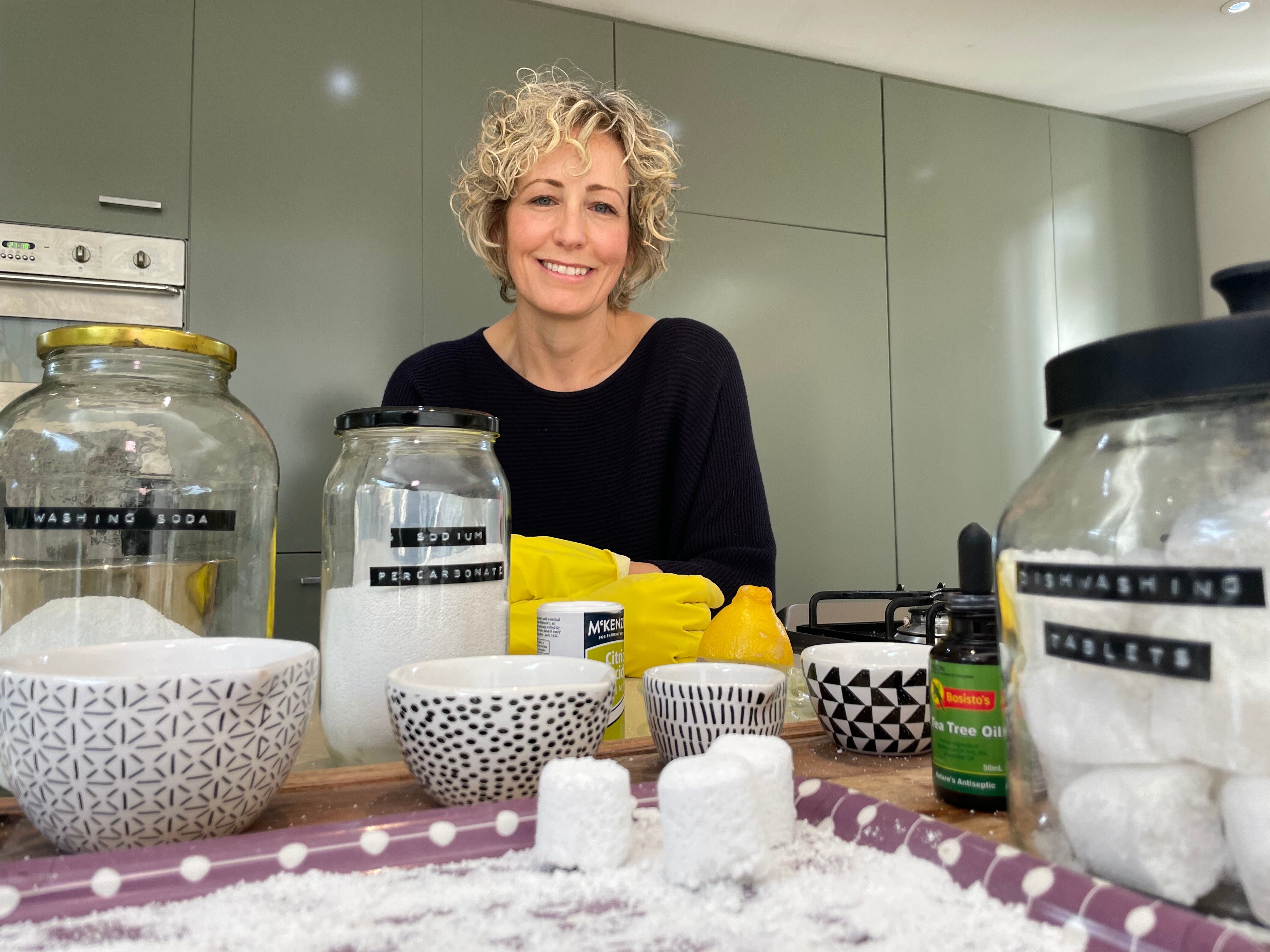 A woman stands at a kitchen table covered in household cleaning products