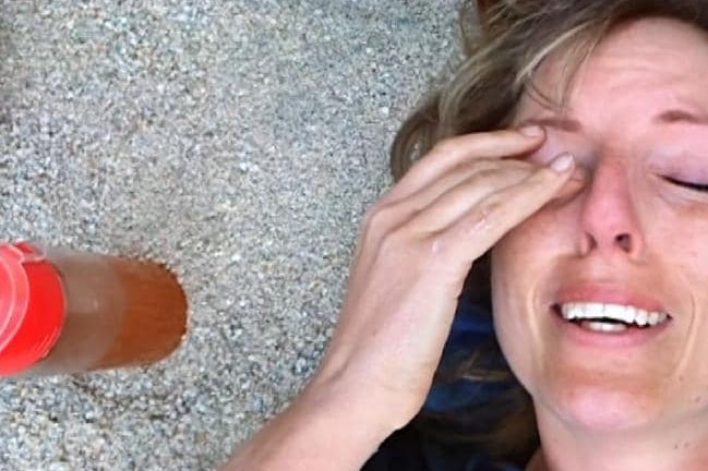 A crying woman is seen rubbing or wiping her eye with a hand while she lays on the ground. Next to her is a drink bottle.