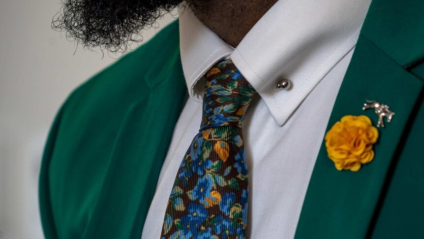 A bearded man wears a crisp white shirt, green blazer and colourful paisley tie