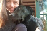 A woman in a grey jumper hugs a black dog to her chin
