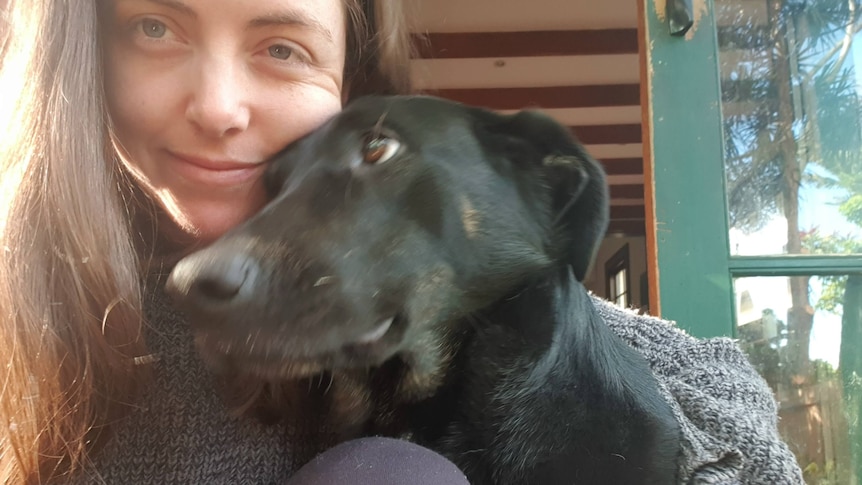 A woman in a grey jumper hugs a black dog to her chin