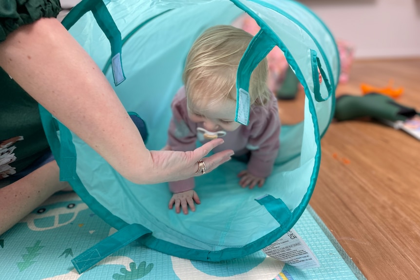 A blonder toddler crawls through a tube. Her mum's arm is keeping it steady.