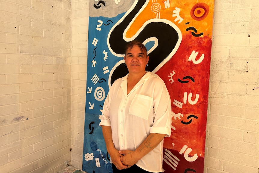 A woman in a white shirt stands in front of a Aboriginal artwork of a snake.