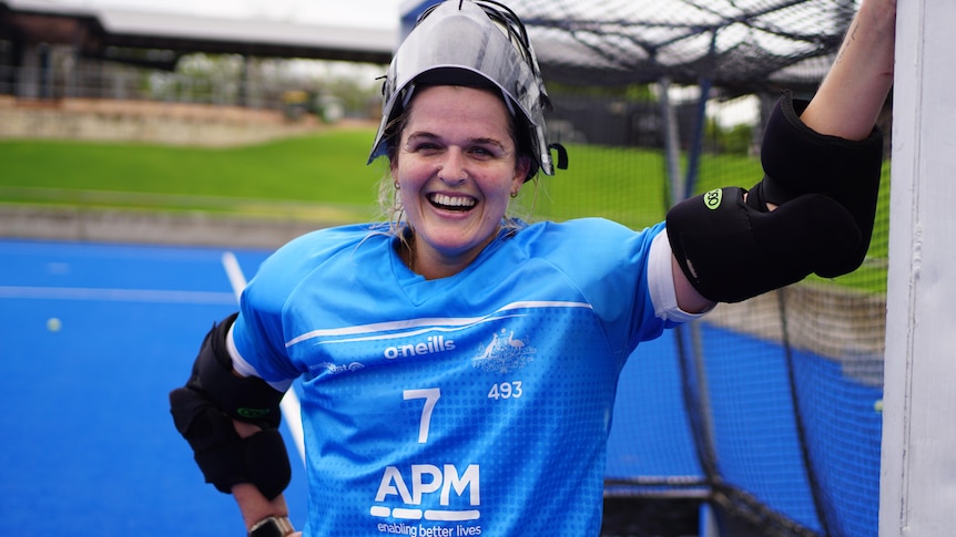 A hockey goalkeeper smiles while leaning against a goalpost