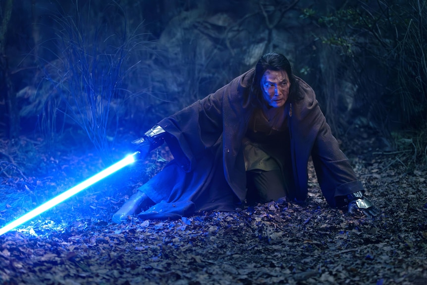 Lee Jung-Jae in a brown cloak with long hair, crouching to the ground holding a blue lightsaber