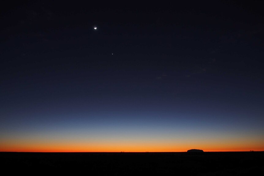 A moon sits in the sky as dusk falls on a desert landscape.