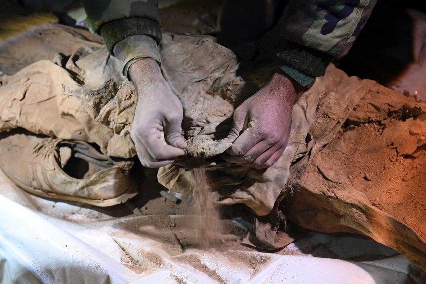 A close-up photo shows hands sifting through the dirt to uncover shoes and human remains.