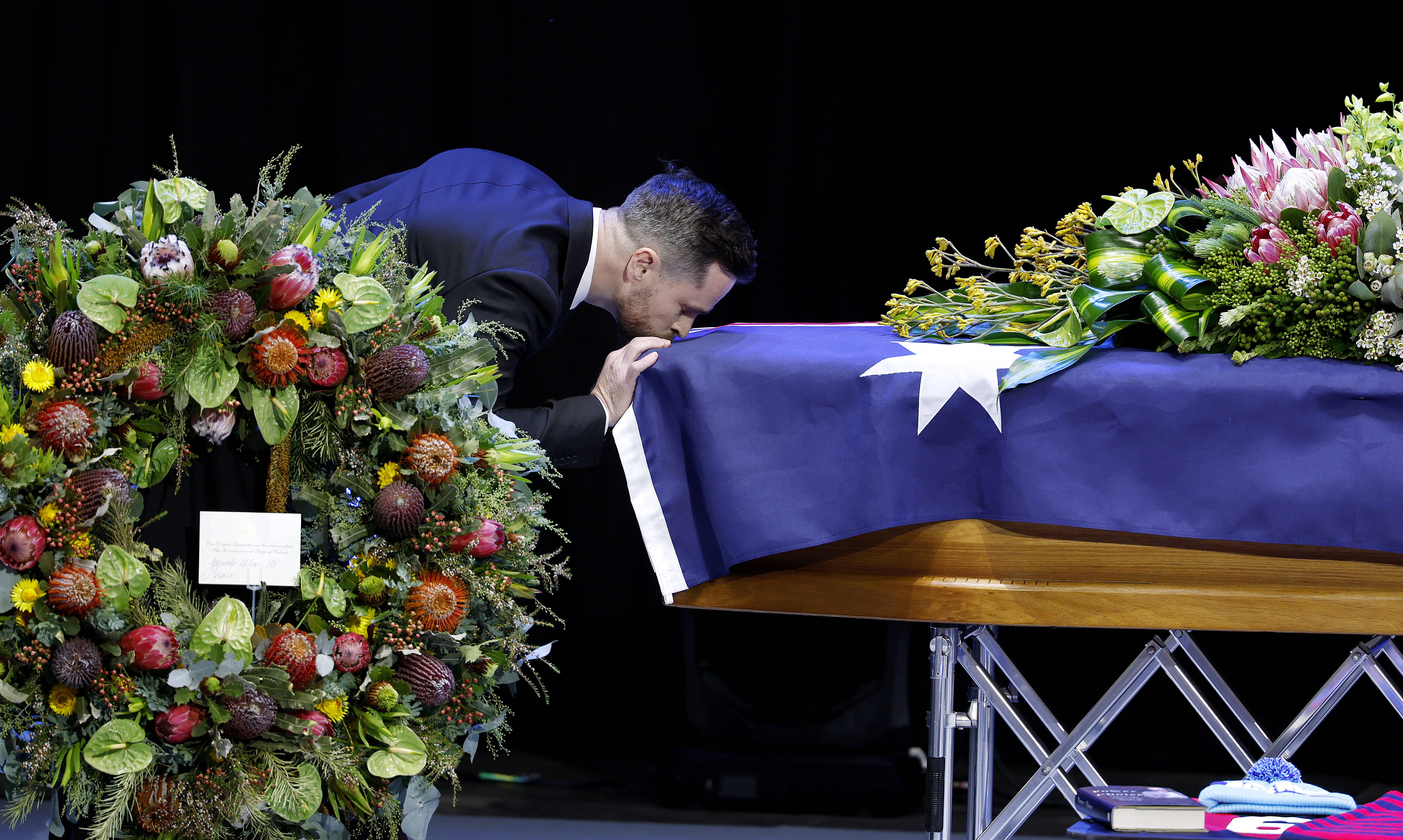 A dark haired man with a beard leans over and kisses a coffin draped in the Australian flag with native flowers on top.