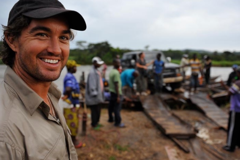 A man grins into the camera while a car is put on a barge