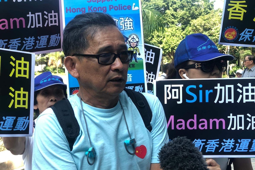 A man in a light blue top with a Chinese flag shaped as a heart speaks into a microphone in front of a crowd.