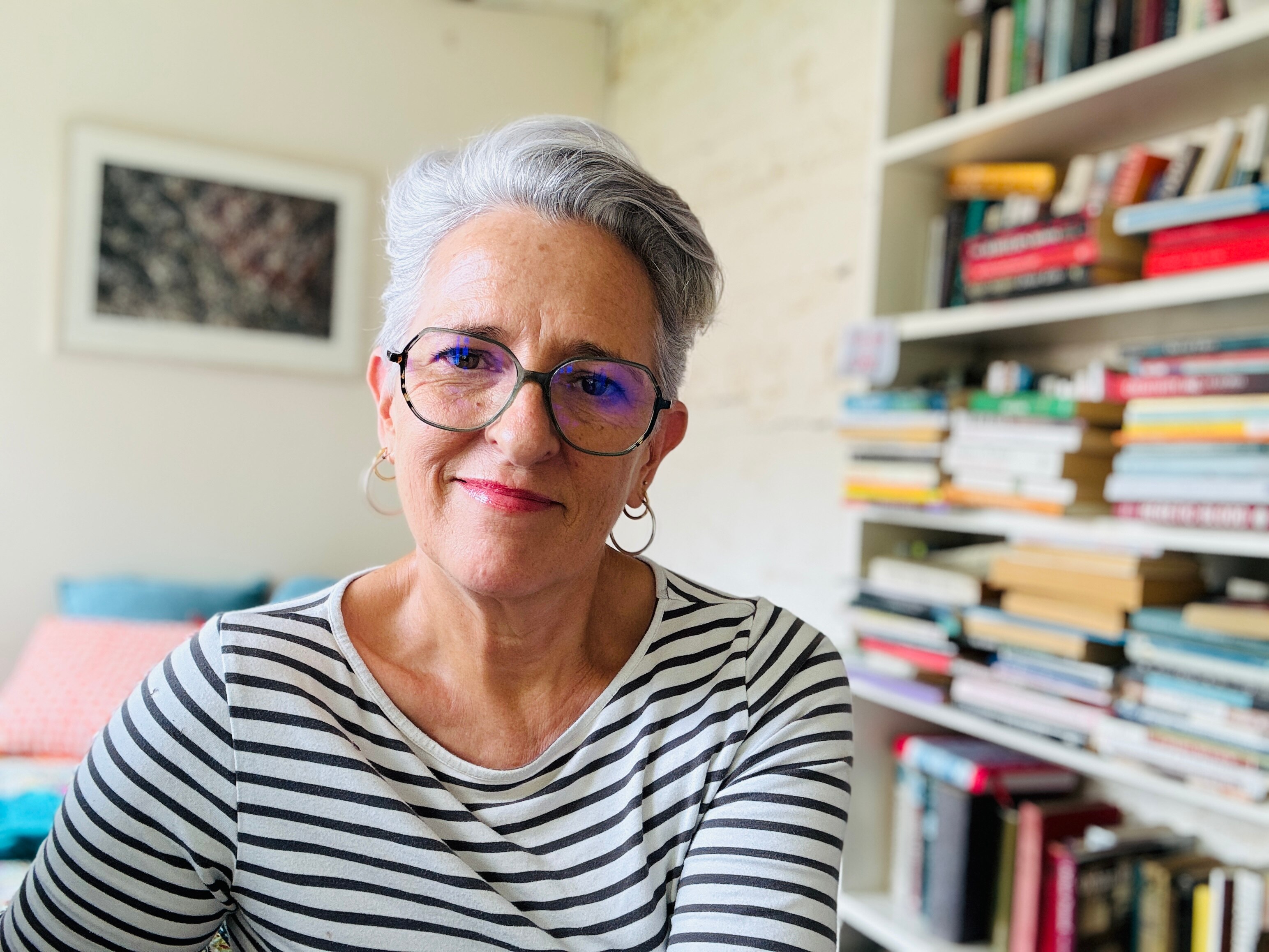 Portrait of author Charlotte Wood, wearing a stripey long sleeve shirt and glasses in her home office, surrounded by books.