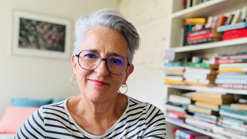 Portrait of author Charlotte Wood, wearing a stripey long sleeve shirt and glasses in her home office, surrounded by books.