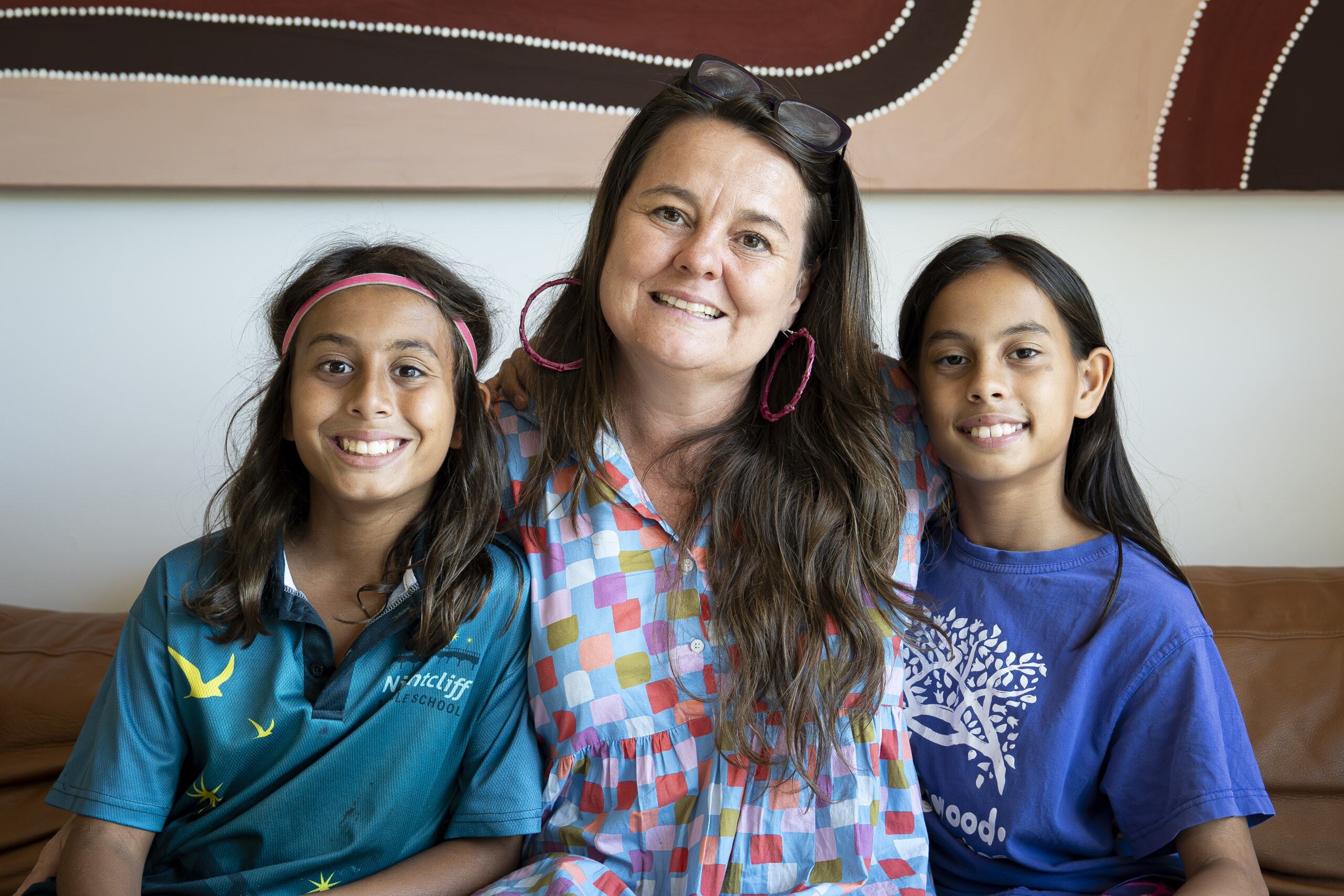 Mother and children together on a brown couch and smile at the camera, with both children leaning into their mother