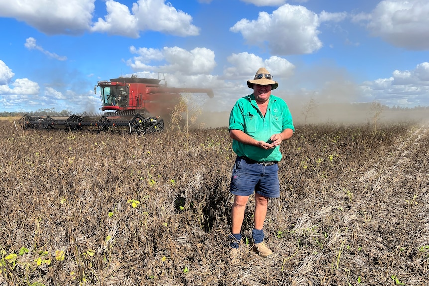 A man in a wide brimmed hat stands in dry looking paddock near a harvester.