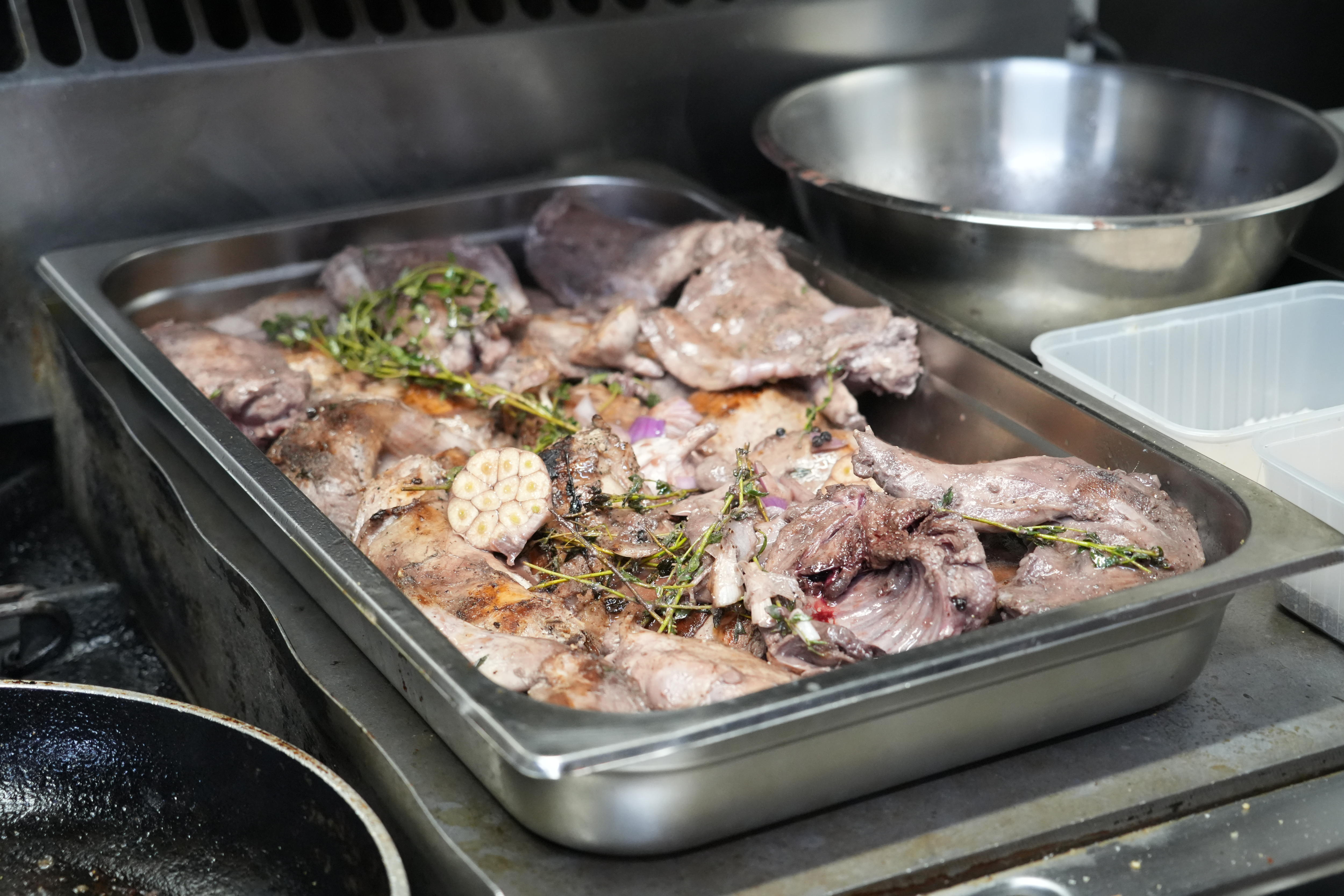 A tray of cooked rabbit meat in a commercial kitchen with onions, garlic and herbs.