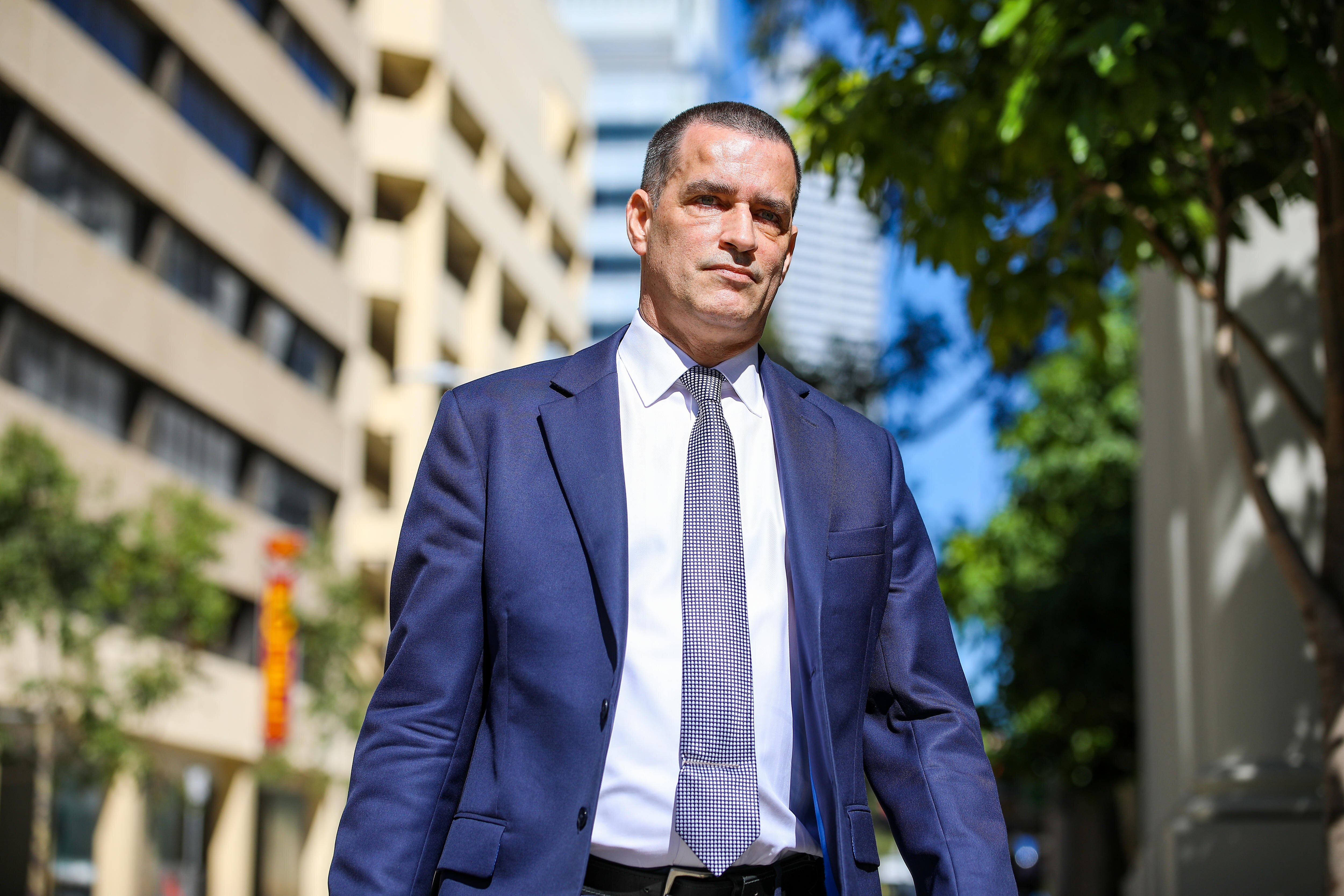 A mid shot of prosecutor Justin Whalley SC walking outside wearin a blue suit, white shirt and blie tie.