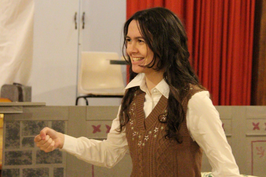 Woman with long brown hair smiles and holds hand up. Wearing brown vest.