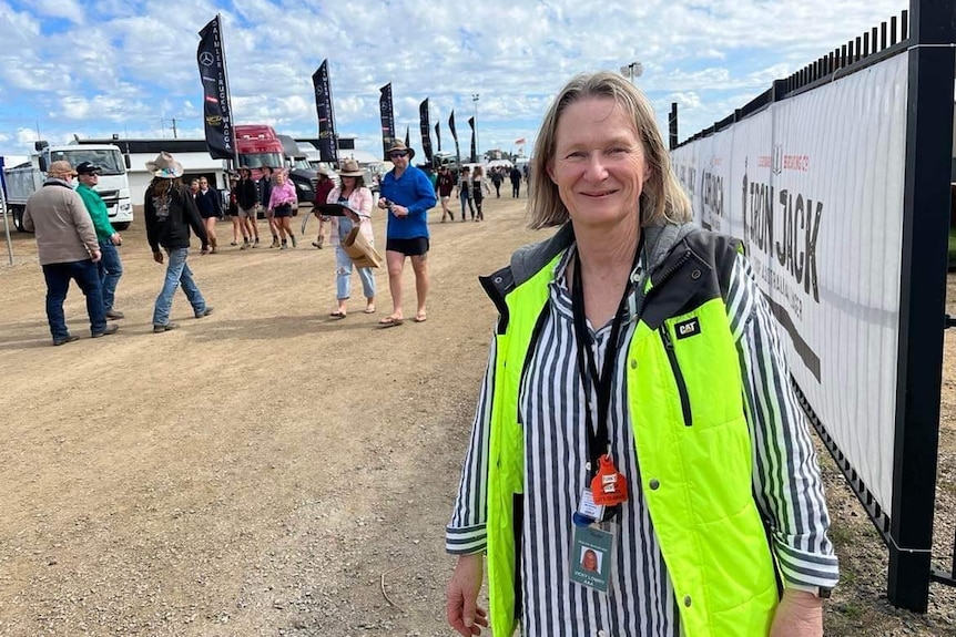 A woman stands on a dirt path, with festival attendees filing past behind her.