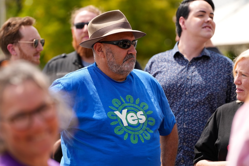 An aboriginal man wearing a hat, sunnies and tshirt with the word 'yes' on it standing in a group of people outside