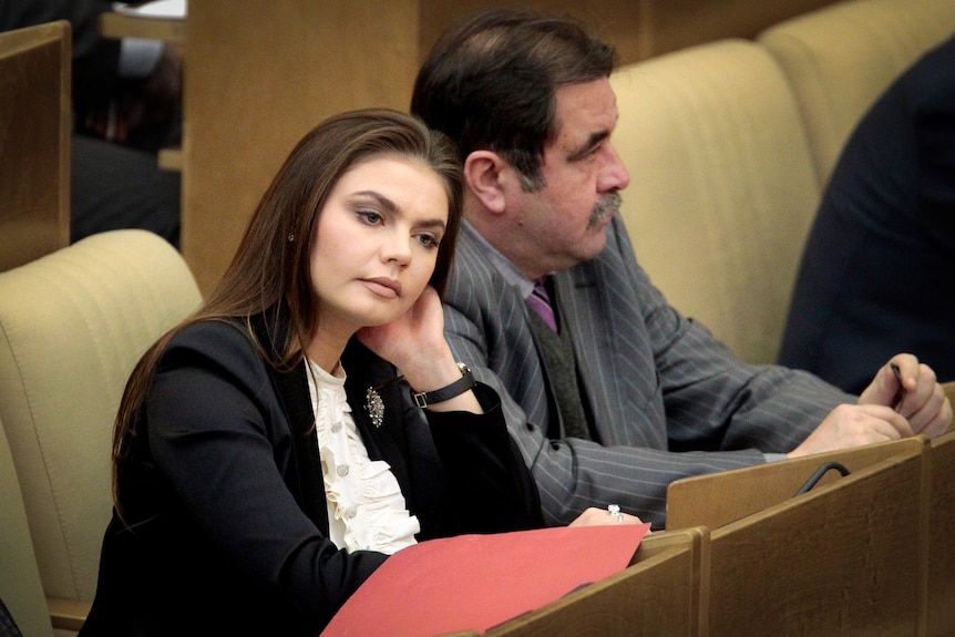 A young woman with dark long hair rests her head on her hand while sitting in a parliamentary setting