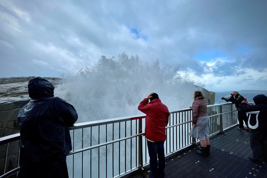 People standing on a viewing platform by the sea being sprayed by a large ocean wave.