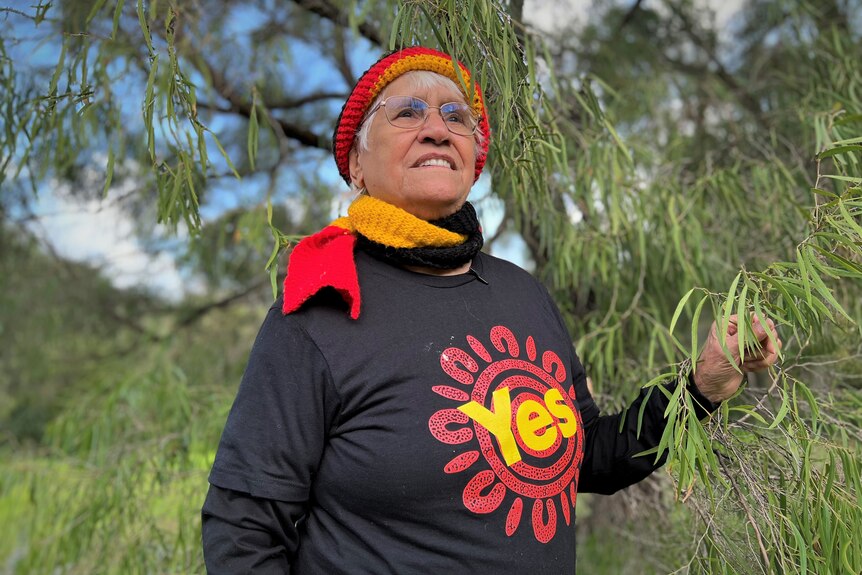 Aboriginal elder woman wearing a shirt that says "yes" in the Aboriginal flag colours standing in the tree