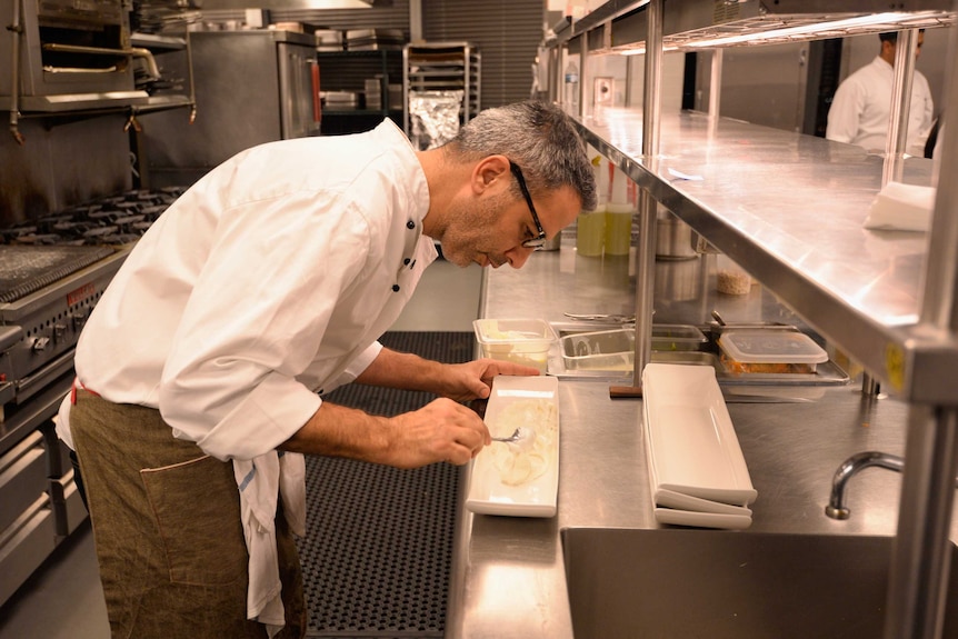 A man wearing a chef's uniform carefully spoons a creamy food onto a rectangular plate in a professional kitchen.