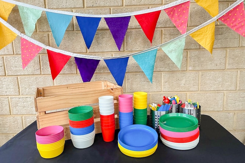 Rainbow plastic plates, cups and bowls are seen in front of a brick wall sitting on a table with rainbow bunting hung overhead.