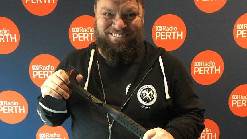 Tyson McMillan stands with an axe in front of the ABC Perth sign.