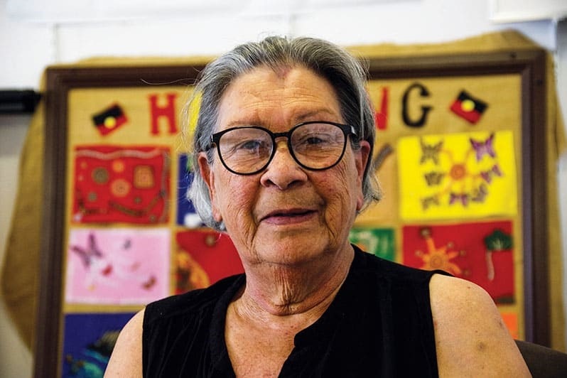 A senior Aboriginal woman wearing spectacles and a sleeveless black top looks into the camera.