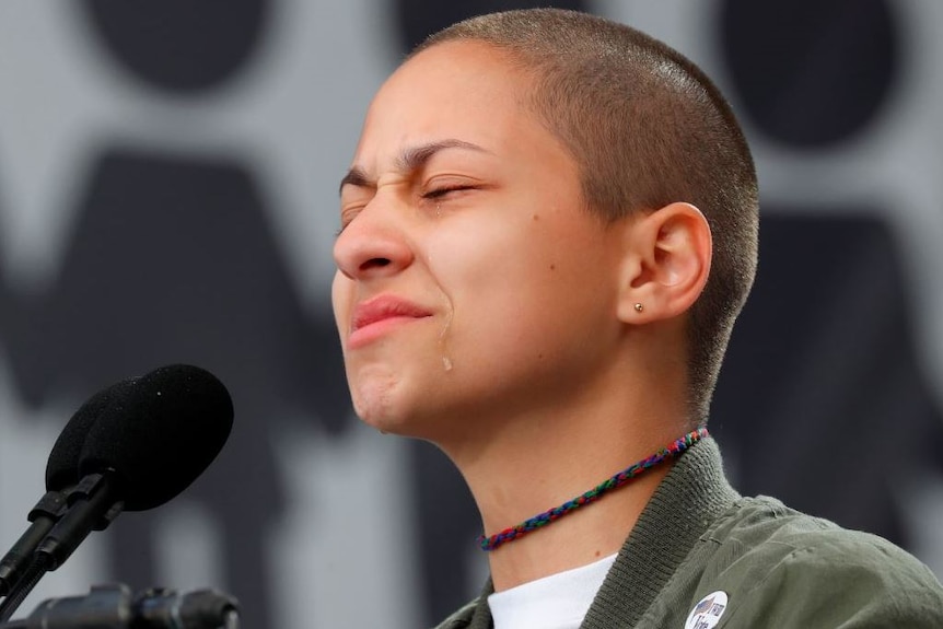 Emma Gonzalez stands behind a microphone and squeezes her eyes shut while a tear runs down her face.
