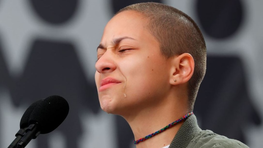 Emma Gonzalez stands behind a microphone and squeezes her eyes shut while a tear runs down her face.