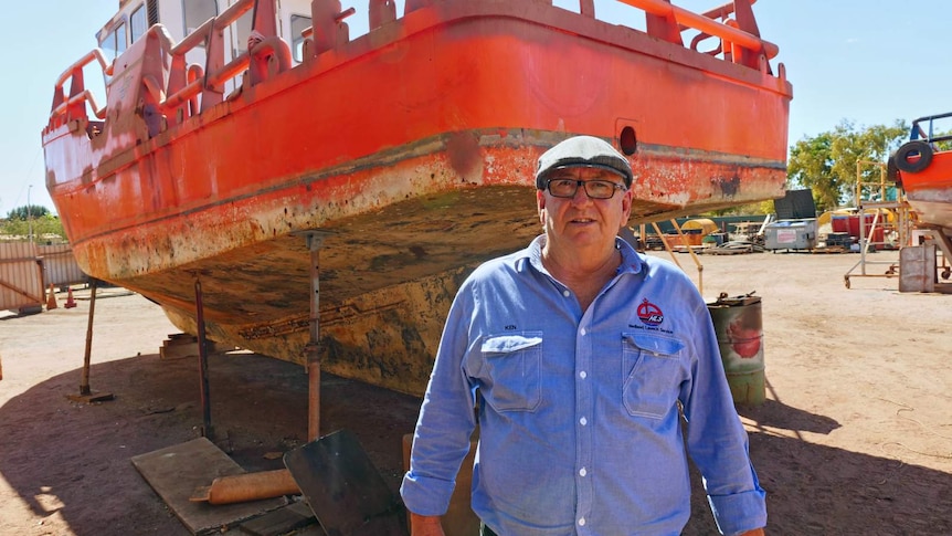 A man wearing a blue shirt, hat and glasses stands in front of a red boat raised out of the water.