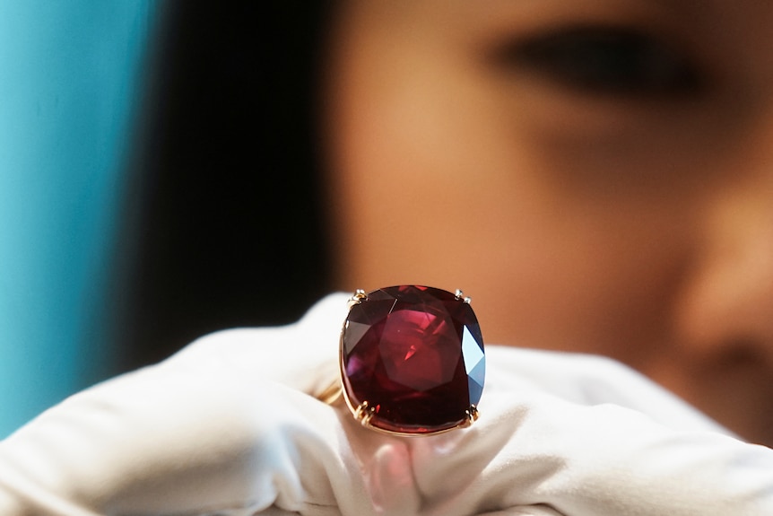 A close up of a gloved hand holding a ruby. A woman's face is blurred in the background.