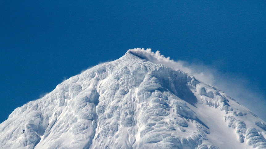 Mountain covered in ice and snow, surrounded by vibrant blue sky, erupts with steam and smoke.