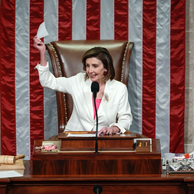 Speaker of the House, Nancy Pelosi in the house chair waving a piece of paper and smiling with the US flag behind her