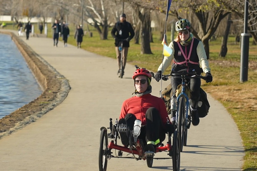 A cyclist follows a person riding a recumbent bicycle on a path along a lake shore.