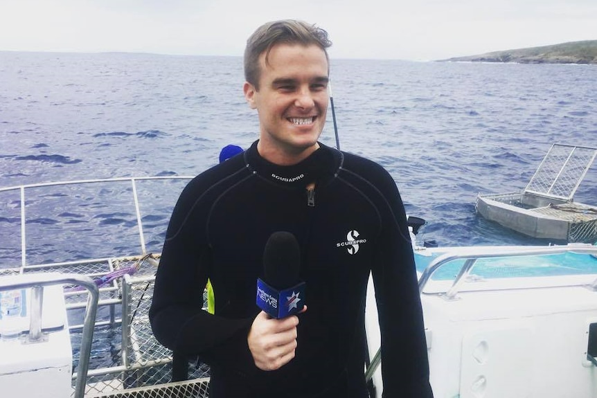 A smiling man in a wetsuit holding a microphone and standing in a boat on water