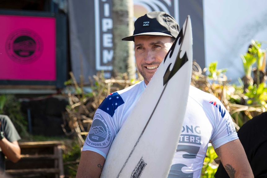 Joel Parkinson wearing a rash vest and hat holds his surfboard on the beach