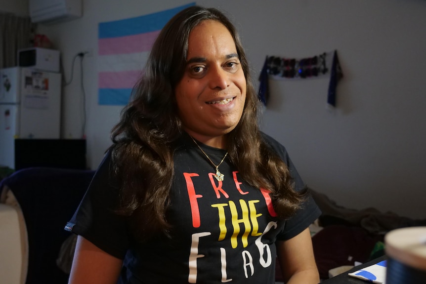 A half-smiling Indigenous woman with long hair, black t-shirt saying 'free the flag', sits in a room.