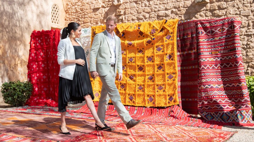 Meghan and Harry walk in front of colourful rugs in Morocco.