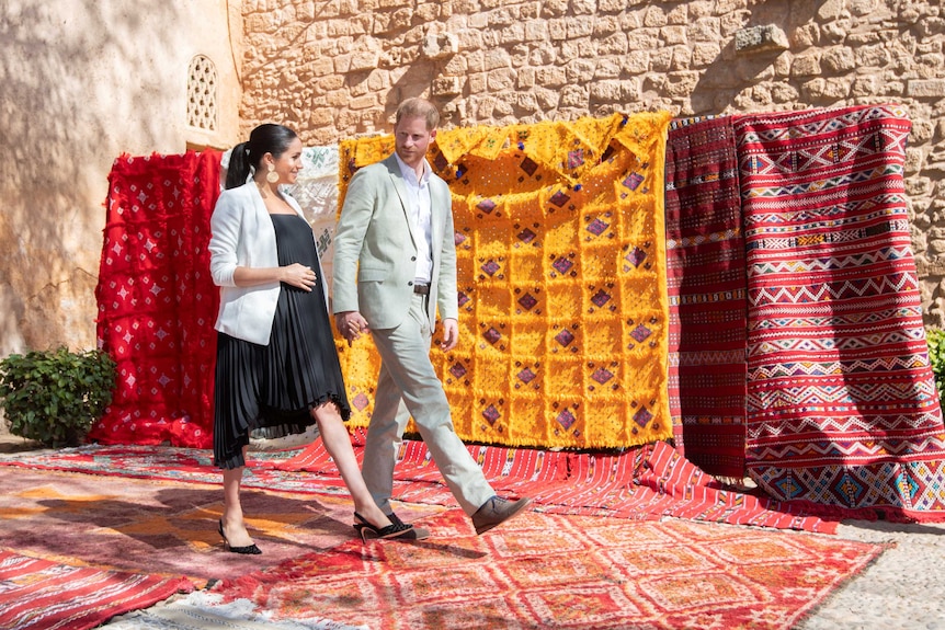 Meghan and Harry walk in front of colourful rugs in Morocco.