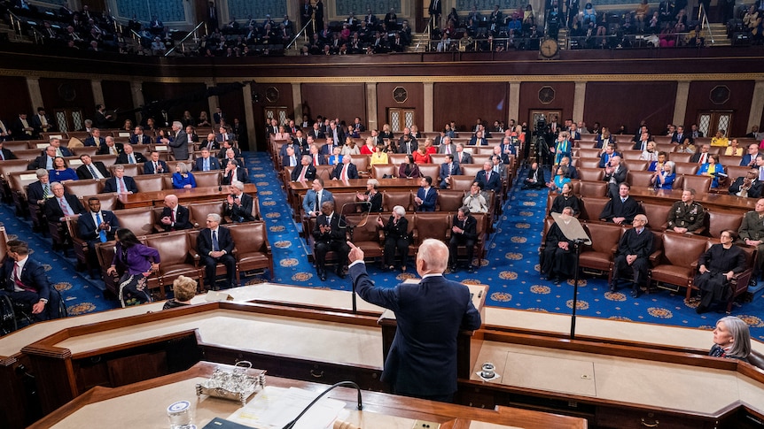 Joe Biden at a lectern surrounded by people in the US House of Representatives chamber