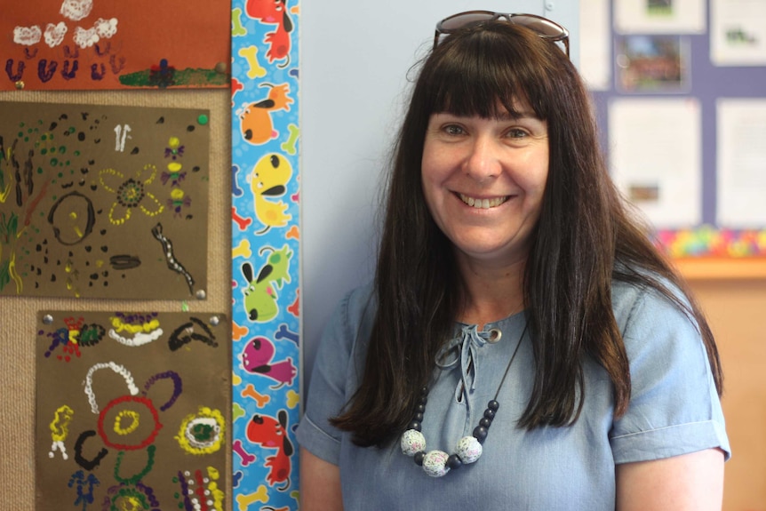 A woman in a blue shirt stands in front of paintings in a primary school classroom.